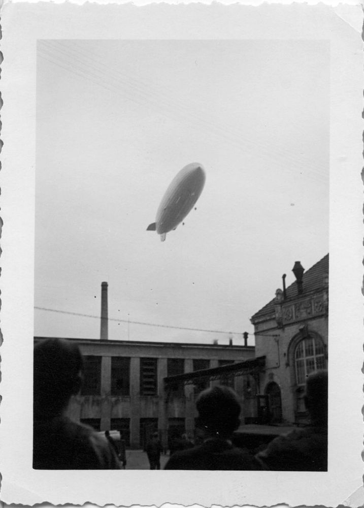 Schwarz-Weiß-Foto von LZ 129 „Hindenburg“ fährt über eine Fabrik in Heidenheim. Im Vordergrund sieht man die Köpfe dreier Personen von hinten. Im Mittelgrund ein Verwaltungsgebäude und die Fabrikhalle mit Schlot.