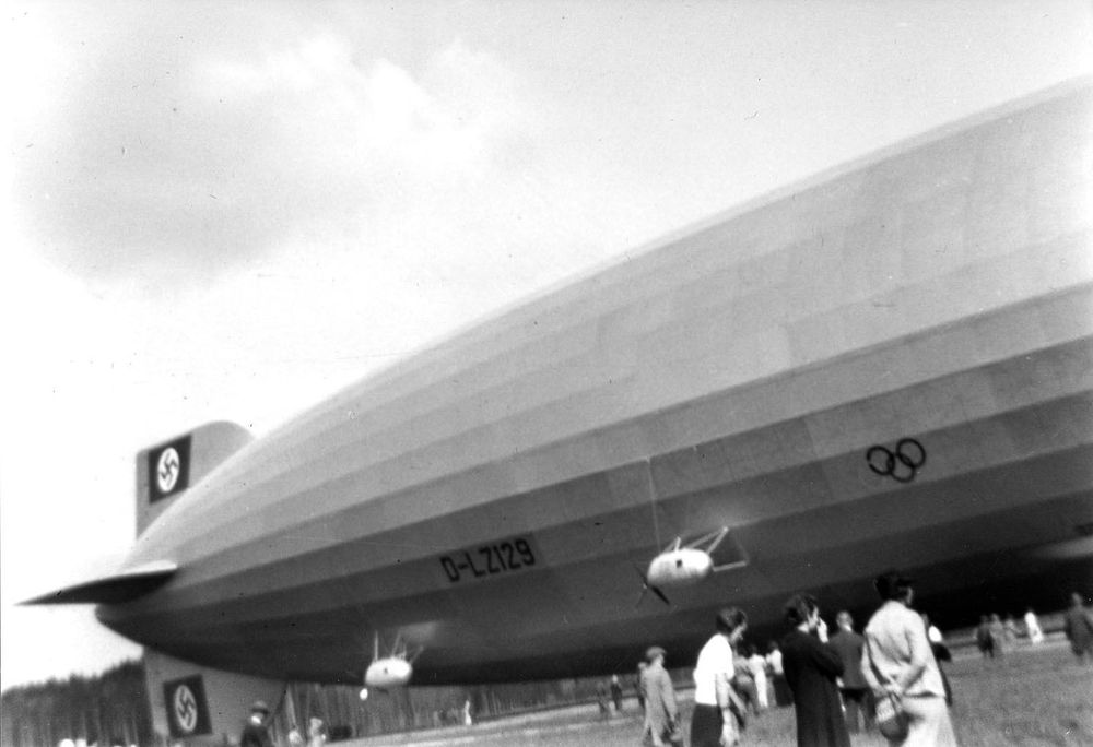 Mittel und Heckansicht des Zeppelins LZ 129 Hindenburg. Der Zeppelin ist am Boden, die Spitze ist nicht zu sehen. Deutlich erkennbar sind die Olympia Ringe und Hakenkreuze. Vor dem Luftschiff stehen einige Menschen.