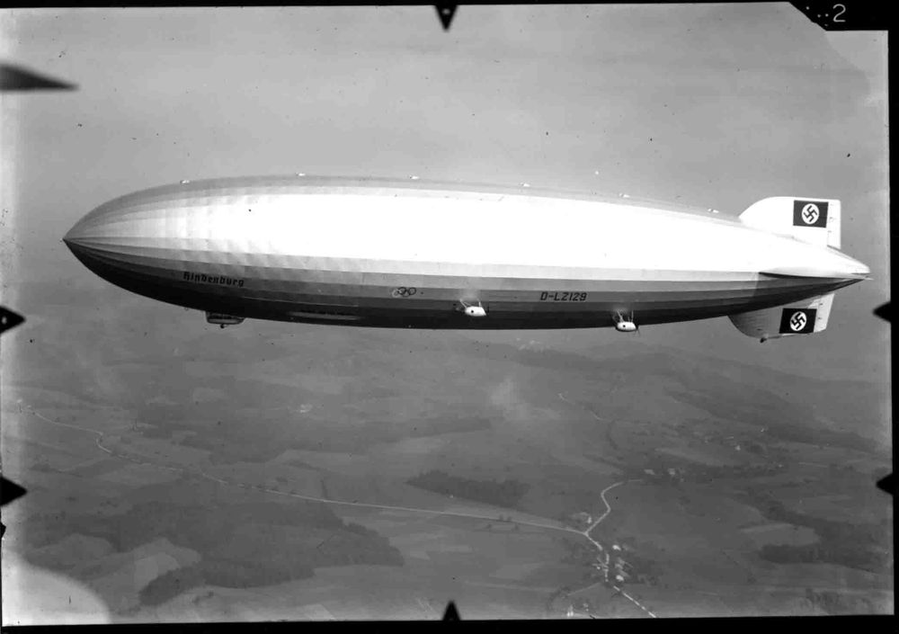 Ansicht auf die Backbordseite des Zeppelins Hindenburg. Hakenkreuze und Beschädigung am Leitwerk sind gut erkennbar. Das Bild wurde aus der Luft aufgenommen. Unter dem Zeppelin ist die Landschaft zu erahnen, fließender Übergang zum Himmel.