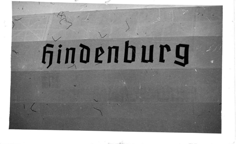 Der Schriftzug von LZ 129 Hindenburg in Frakturschrift. Er ist mittig auf dem Foto zu sehen, ansonsten sieht man nur die umlaufende graue Hülle. Durch den Ausschnitt ist der Zeppelin ansonsten nicht erkennbar. 