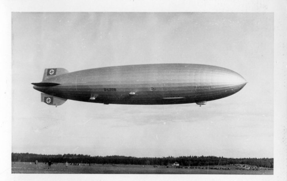 Der Zeppelin LZ 129 fliegt am Himmel von links nach rechts. Unter ihm ist Wiese und im Hintergrund Wald zu sehen. Auf den Seitenleitwerken sind die Hakenkreuze und eine Beschädigung erkennbar. 