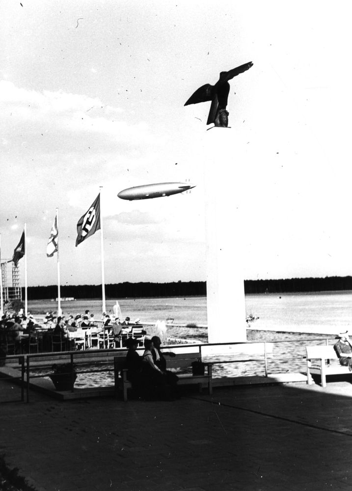Zentral im Hintergrund ist der fahrende Zeppelin LZ 130 am Himmel zu sehen. Im Vordergrund der Flughafen Frankfurt. Links weht die Hakenkreuzflagge, recht vom Zeppelin steht die Ikarus Säule.