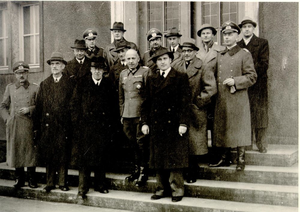 Eine Gruppe teils uniformierter Männer stehen vor einem Gebäude. Darunter auch die in zivil gekleideten Hugo Eckener, Ludwig Dürr und Wernher von Braun sowie Vertreter des Militärs. Die Gruppe besteht aus 14 Personen. 