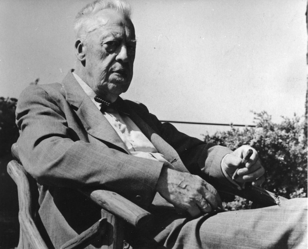 Hugo Eckener im Jahr 1954 im Alter von 86. Er sitzt in einem Gartenstuhl unter freiem Himmel. In seiner linken Hand hält er eine Zigarre.