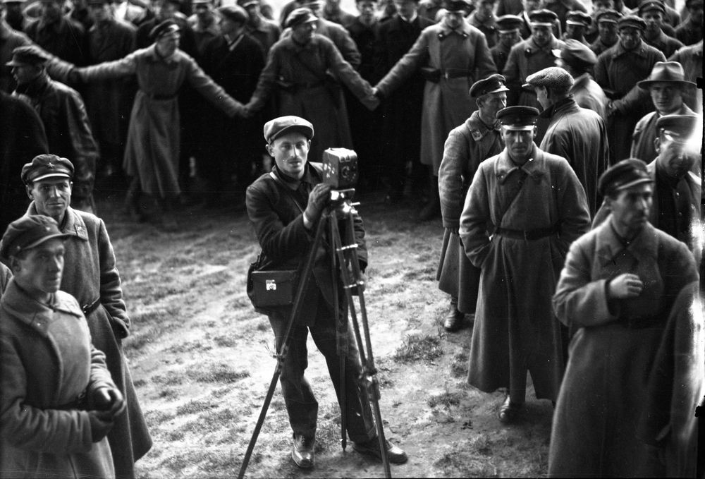 Ein Kameramann von vorne gesehen mit Kamera auf dem Stativ, umringt von den Haltemannschaften. Im Hintergrund bilden russische Soldaten eine Absperrkette.