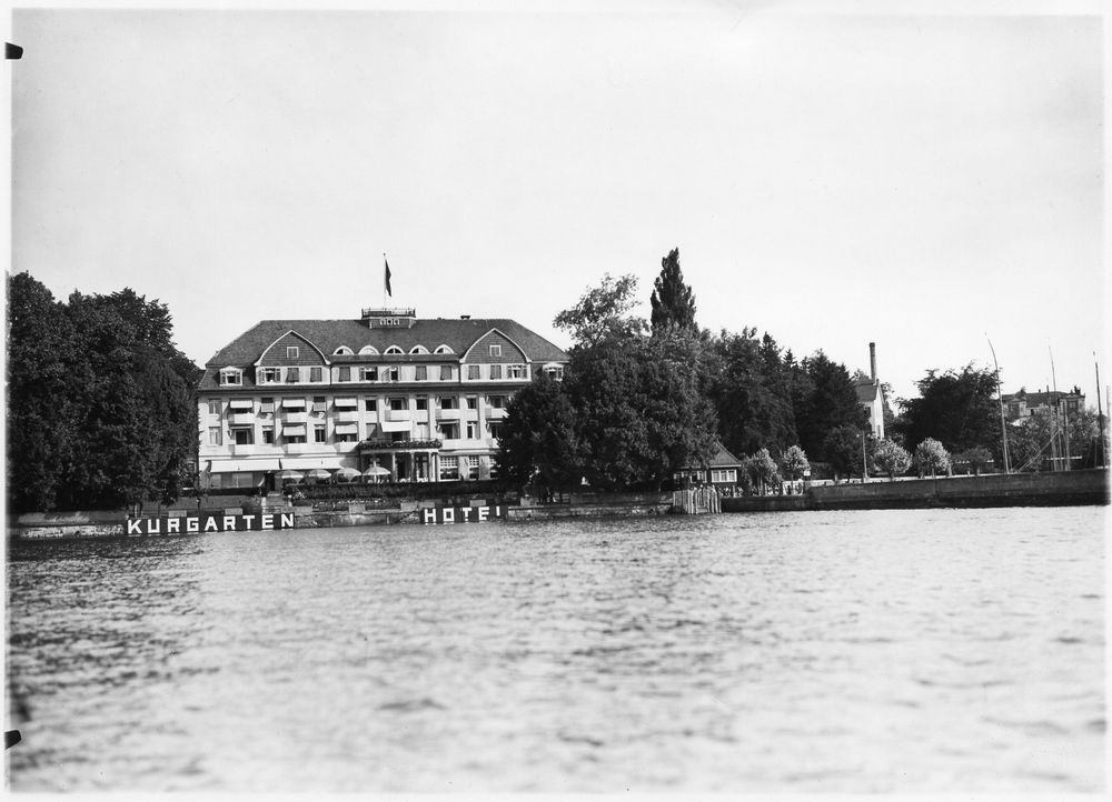 Das Kurgartenhotel in Friedrichshafen, wo die Jubiläumsfeier stattfand. Blick vom See aus auf die Kaimauer und das Gebäude. Rechts davon ist ein Steg, neben dem Hotel sind noch weitere Gebäude zu sehen. 