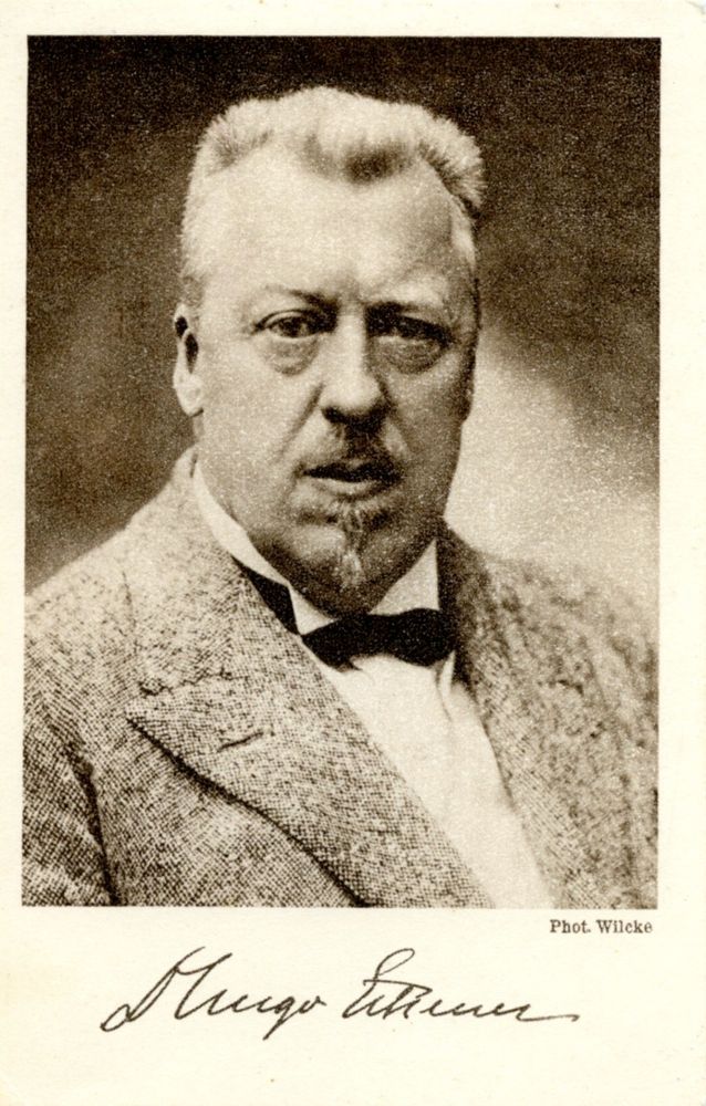 Postkarte mit dem Porträt von Hugo Eckener mit Unterschrift. Eckener trägt ein weißes Hemd mit einem melierten Anzug und dunkler Fliege, außerdem einen Ziegenbart und Bürstenhaarschnitt. Sein Kopf ist rechteckig und er hat markante Tränensäcke. 