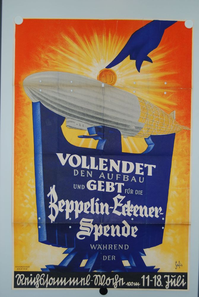Auf einem Plakat befindet sich auf einem stilisierten blauen Adler der Text: „Vollendet den Aufbau und gebt für die Zeppelin-Eckener-Spende“. Über dem Kopf und den ausgebreiteten Schwingen fliegt ein halbfertiger, wie eine Spardose gestalteter Zeppelin. 