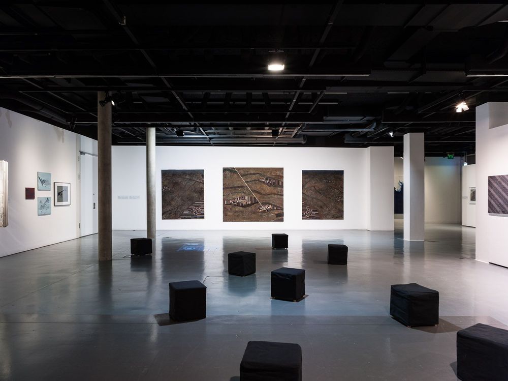 In dem Ausstellungsraum befinden sich schwarze, quadratische Hocker auf dem Boden. An der weißen Wand dahinter und an den Wänden rechts und links hängen verschiedene Bilder.