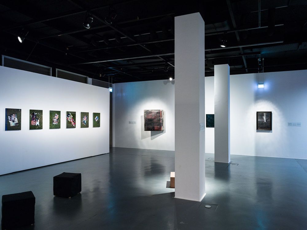 In dem Ausstellungsraum befinden sich schwarze, quadratische Hocker auf dem Boden. An den weißen Wänden dahinter hängen verschiedene Bilder.