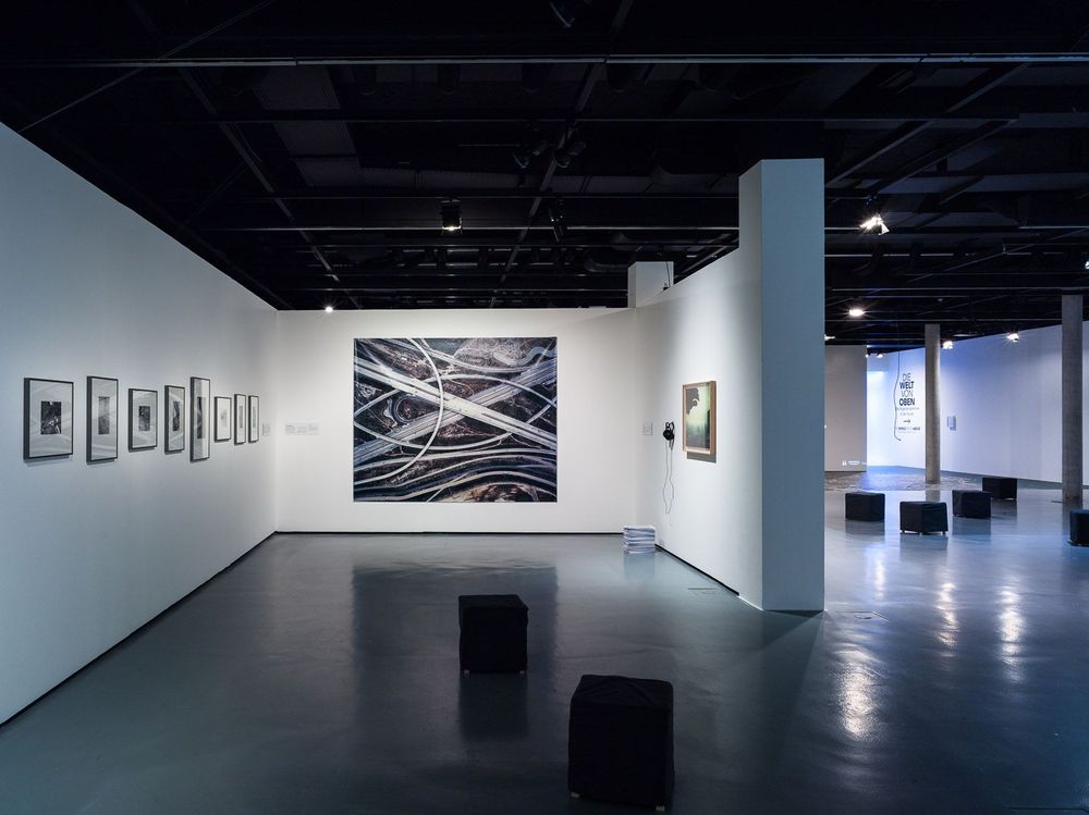 In dem Ausstellungsraum befinden sich schwarze, quadratische Hocker auf dem Boden. An der weißen Wand dahinter und an der Wand rechts hängen verschiedene Bilder.