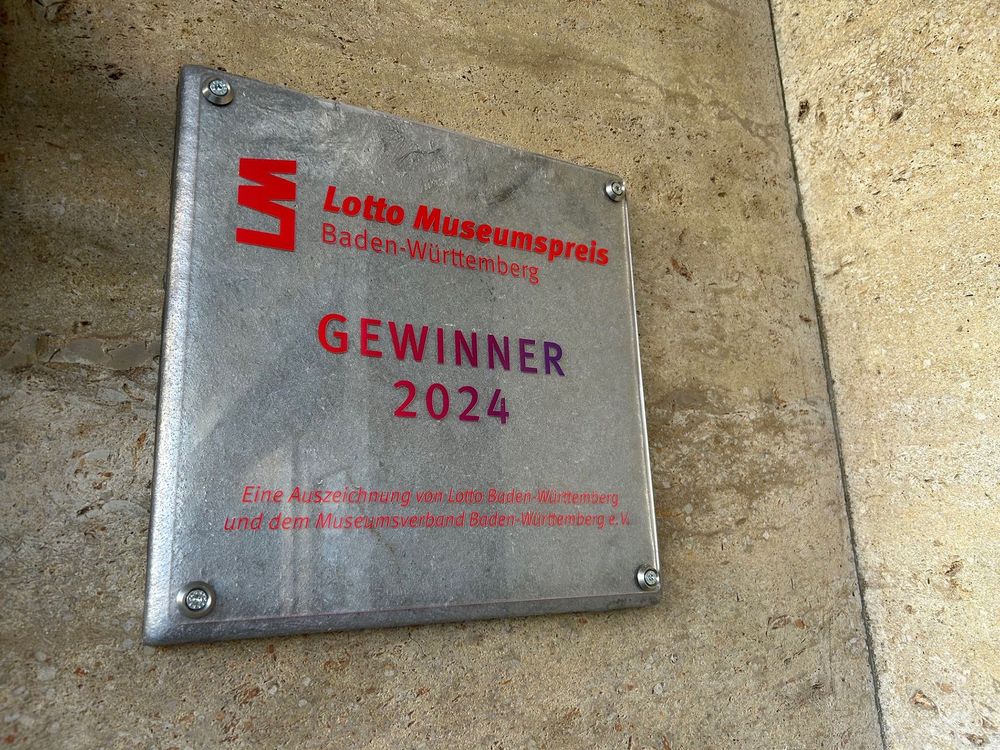 Bild bei Tageslicht außen einer quadratischen Metallplatte mit roter Aufschrift auf einer hellen Steinwand
