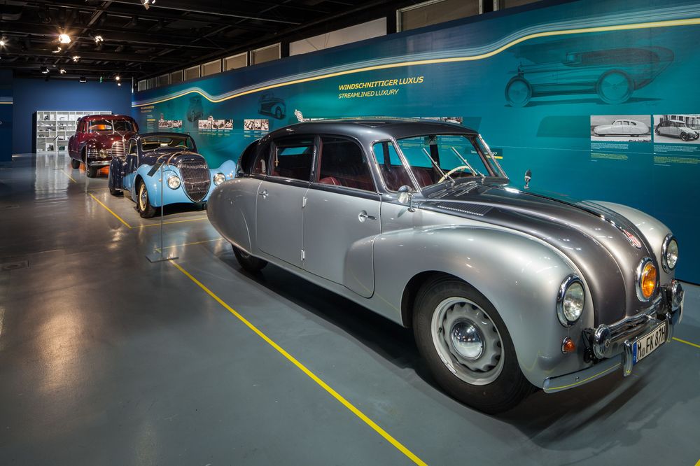 Die Ansicht zeigt einen silbernen Oldtimer von der Seite. Das Auto hat vier Türen, mit je zwei Sitzbänken.