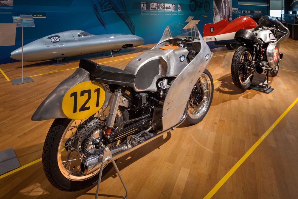 Die Ansicht zeigt zwei ältere Renn-Motorräder. Bei dem Vorderen ist über der Schutzklappe des Hinterrades ein gelber Kreis mit der Nummer 121 aufgedruckt. 