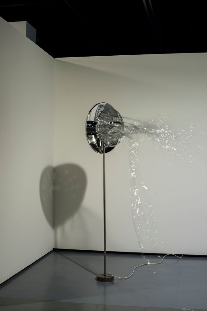 Vor einer weißen Wand steht ei silberfarbener Ventilator, der silberne Schnüre wegbläst.