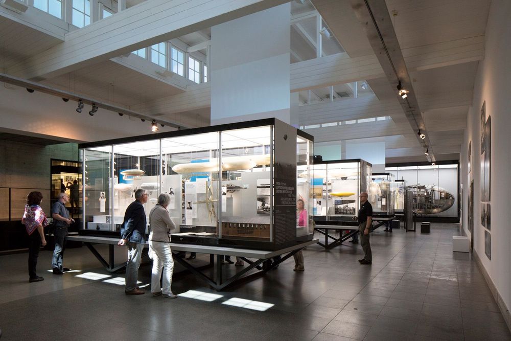 Eine lichtdurchflutete Ausstellungsfläche mit mehreren Informationstafeln und großen Glasvitrinen, in denen sich Objekte befinden. Mehrere Personen schauen sich die Tafeln und Vitrinen interessiert an.