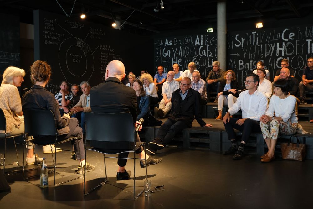 Menschen sitzen auf einem Palettenhügel in der Ausstellung "Into the deep. Minen der Zukunft" und hören den sprechenden Personen im Vordergrund gespannt zu.