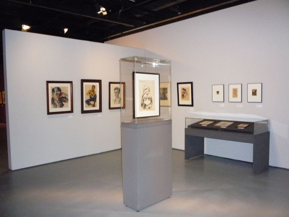 Die Ausstellungsansicht zu "Elfriede Lohse-Wächtler. Ein Leben zwischen Kunst und Verzweiflung" zeigt eine Ausstellungsfläche mit einer Vitrine in der Mitte und verschiedenen Bilderrahmen an den Wänden. 
