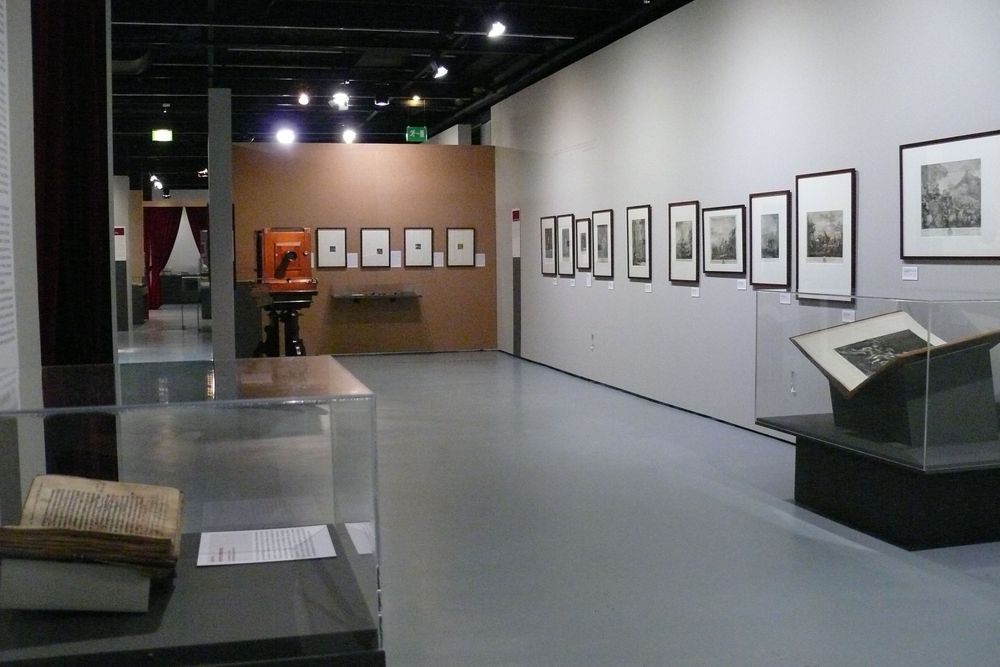 Die Ausstellungsansicht zu "Gestochen scharf" zeigt zwei Vitrinen, in denen sehr alte und große Bücher aufgeschlagen liegen. Im Hintergrund an den Wänden hängen verschiedene Bilder.