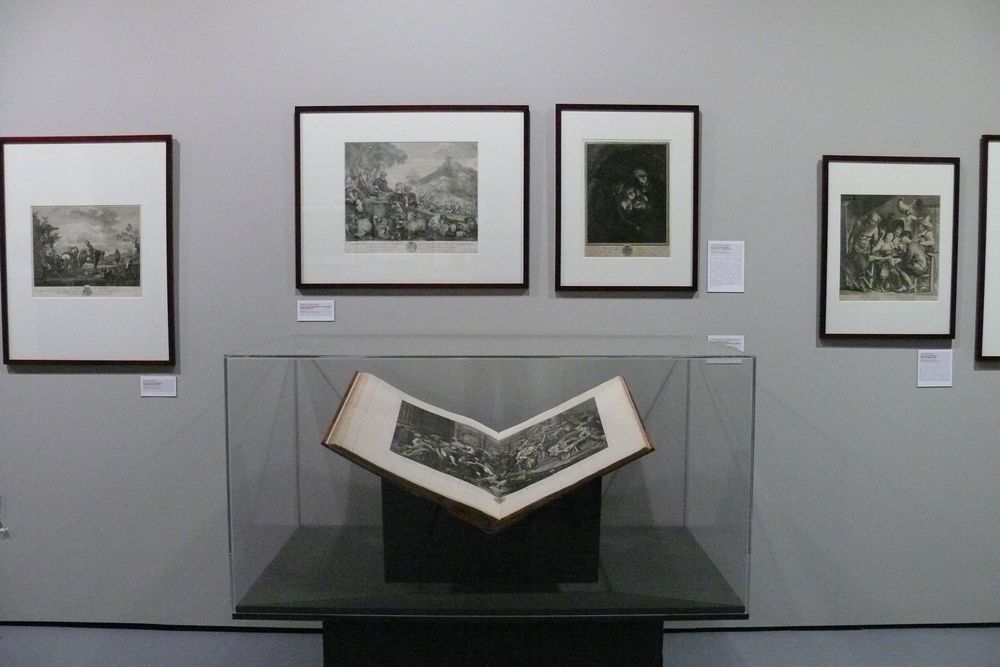 Die Ausstellungsansicht zu "Gestochen scharf" zeigt frontal eine Vitrine in der ein aufgeschlagenes, großes Buch liegt. Direkt dahinter an der Wand hängen verschiedene Bilder.