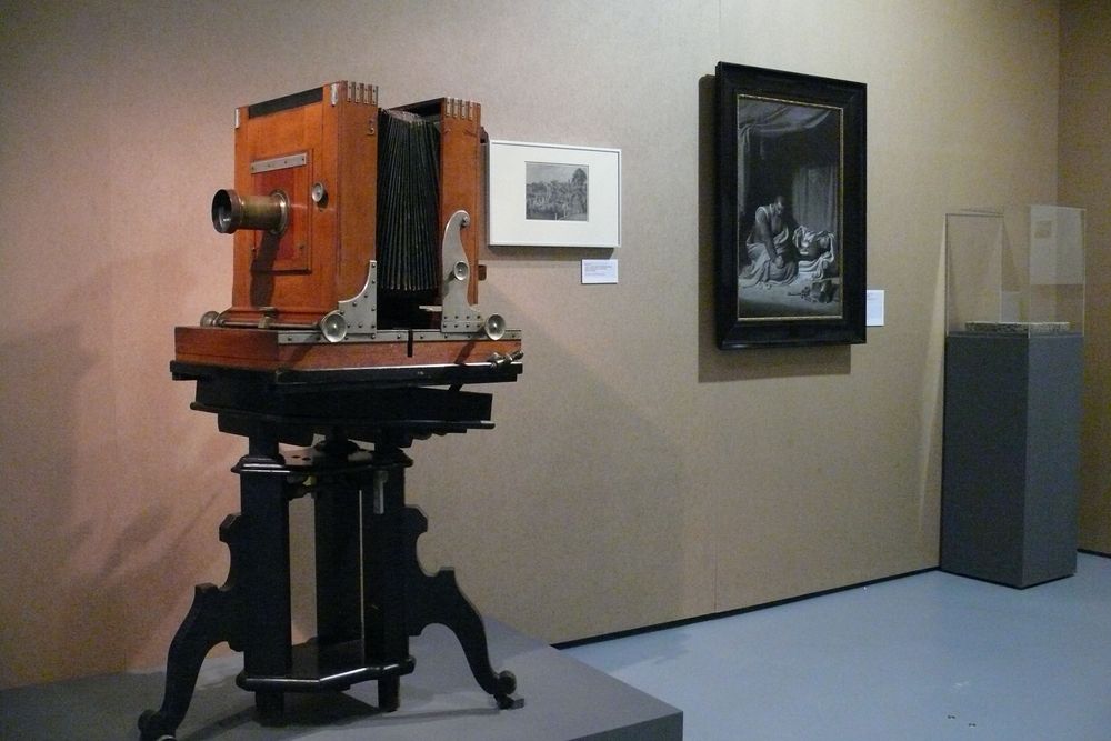 Die Ausstellungsansicht zu "Gestochen scharf" zeigt eine alte Kamera, die auf einem Podest steht, im Hintergrund steht eine kleinere Vitrine und an der Wand hängen zwei unterschiedliche Bilderrahmen.