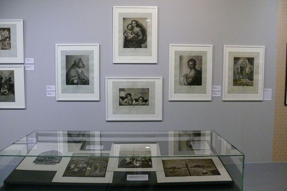 Die Ausstellungsansicht zu "Gestochen scharf" zeigt eine Vitrine in der sich vier Bilder befinden. Dahinter an der Wand hängen weitere Bilder in Rahmen. 