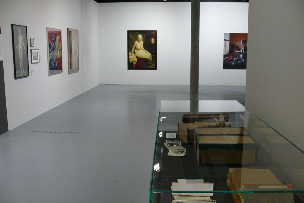 Die Ausstellungsansicht zu "Gestochen scharf" zeigt einen großen leeren Raum an dem ein paar Bilderrahmen an den Wänden hängen. Im Vordergrund des Flurs, der in den Raum führt steht eine Vitrine mit Exponaten.