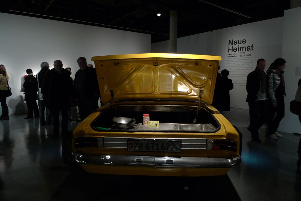 Die Ausstellungsansicht von "Neue Heimat" zeigt ein gelbes Auto mit geöffneter Heckklappe. Im Hintergrund bewegen sich Menschen um das Auto.
