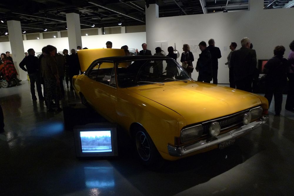 Die Ausstellungsansicht von "Neue Heimat" zeigt ein gelbes Auto mit geöffneter Heckklappe. Neben einem der Reifen steht ein kleiner Fernseher, auf dem ein Video abspielt. Um das Auto herum stehen Besucher*innen der Ausstellung.