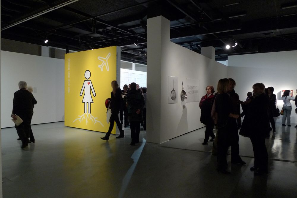 Die Ausstellungsansicht von "Neue Heimat" zeigt zwei voneinander abgetrennte Ausstellungsräume und Besucher*innen, die sich durch die Räume bewegen.