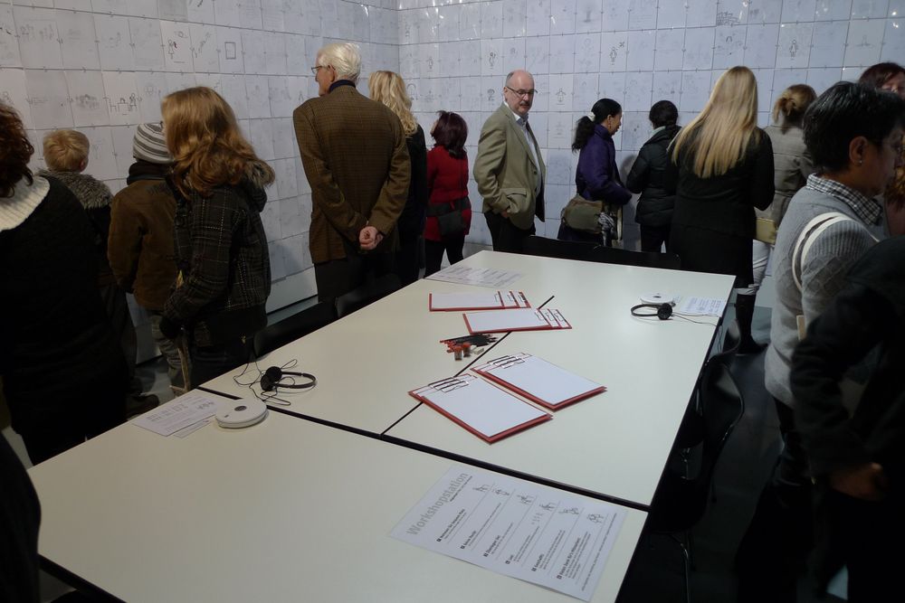 Die Ausstellungsansicht von "Neue Heimat" zeigt drei zusammengeschobene Tische auf denen Blätter und Kopfhörer liegen. Um die Tische herum stehen Besucher*innen, die Bilder und Texte an den Wänden anschauen.