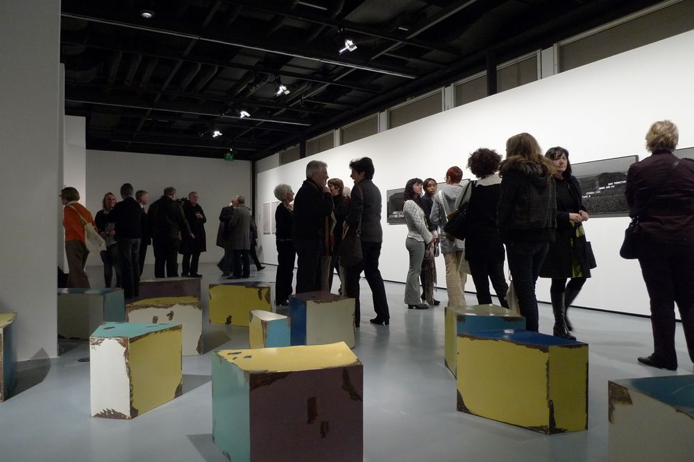 Die Ausstellungsansicht von "Neue Heimat" zeigt mehrere bunte und eckige Objekte, die unterschiedlich verteilt auf dem Boden stehen. Dahinter unterhalten sich verschiedene Besucher*innen-Gruppen.
