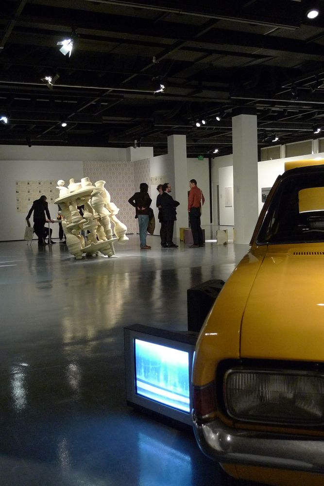 Die Ausstellungsansicht von "Neue Heimat" zeigt ein gelbes Auto, neben dessen Reifen ein kleiner Fernseher steht, auf dem ein Video abspielt. Im Hintergrund stehen Besucher*innen um ein weiteres großes Objekt herum.