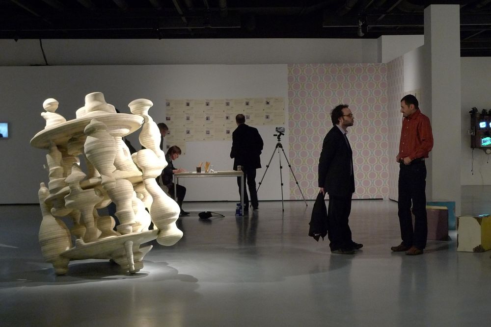 Die Ausstellungsansicht von "Neue Heimat" zeigt ein großes Objekt, das an einen Kreisel erinnert, welches mitten im Raum steht. Im Hintergrund sind Besucher*innen zu sehen.