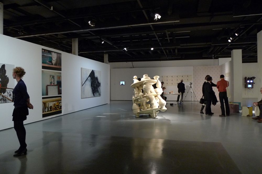 Die Ausstellungsansicht von "Neue Heimat" zeigt ein großes Objekt, das an einen Kreisel erinnert, welches mitten im Raum steht. Im Hintergrund sind Besucher*innen zu sehen.