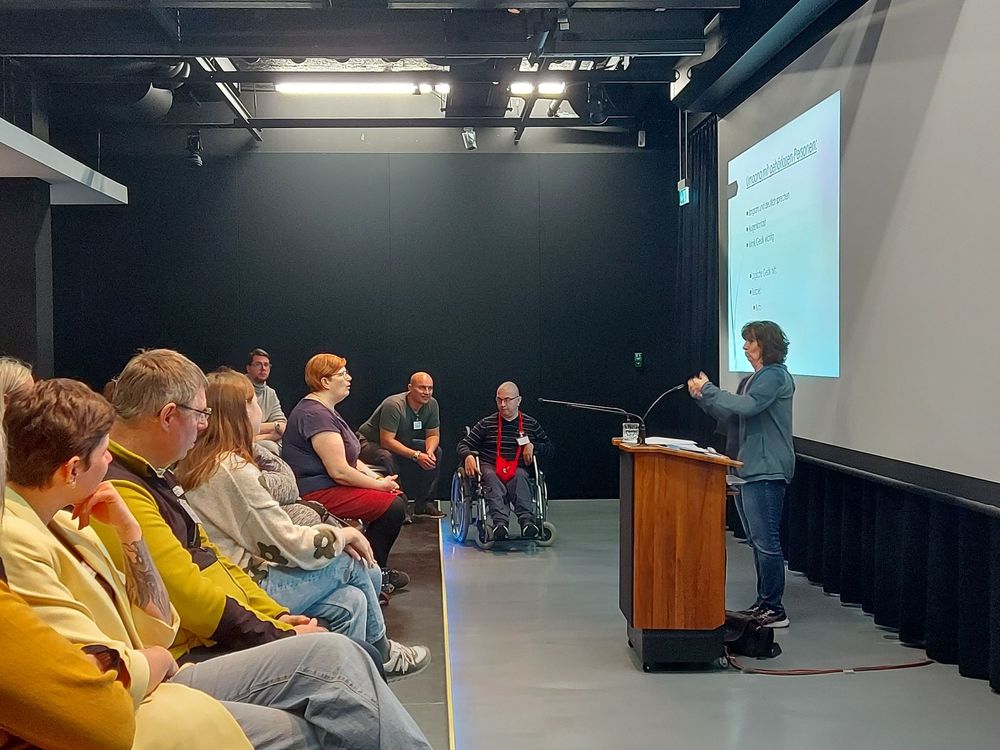 Rechts im Bild steht Petra Ehrmann, die Gebärdensprachdozentin, am Rednerpult und gebärdet. Sie befindet sich im Medienraum des Zeppelin Museums. Links im Bild sitzen die Teilnehmer*innen auf Stühlen und Rollstühlen.