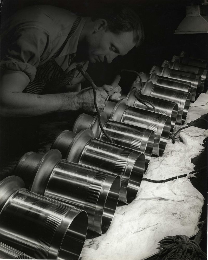 Die schwarz-weiß Fotografie zeigt einen Mann, der mit einer Maschine an einem Objekt arbeitet.