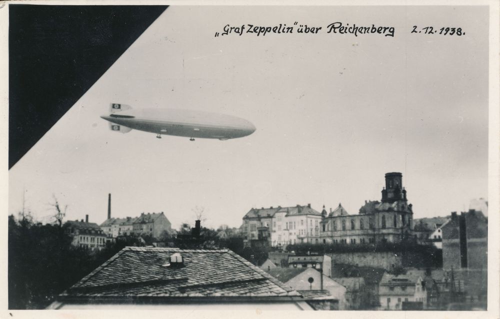 Postkarte mit Zeppelin, wohl aus einem Fenster gemacht. Die linke obere Ecke ist daher schwarz. Unter dem Zeppelin liegt die Stadt Reichenberg. Im rechten Teil ist die Synagoge zu sehen. Ihr fehlen die Kuppel und der Dachstuhl, die verbrannt wurden.