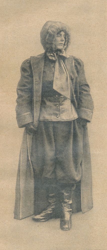 Die historische Aufnahme zeigt eine Frau in damals üblicher Kleidung für Luftschifferinnen. Sie trägt flache Stiefel, einen dicken, sehr langen Mantel, sehr weite Hosen und eine dicke Kapuze.