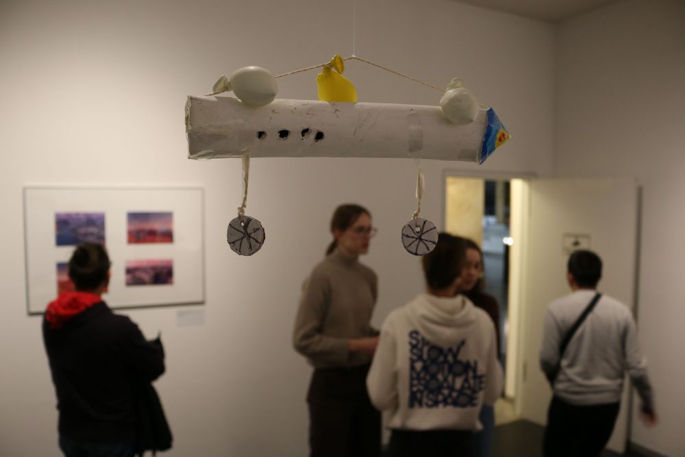 Im Hintergrund: Mehrere Menschen unterhalten sich und begutachten Bilder. Im Vordergrund: Ein aus einer Küchenrolle gebasteltes Luftschiff.