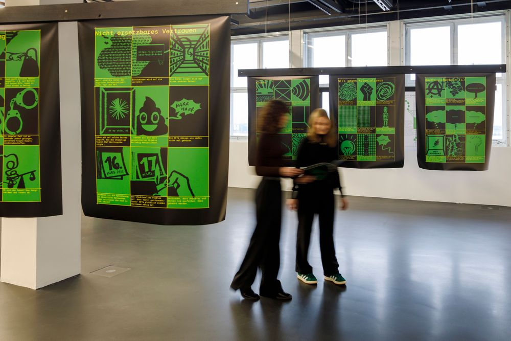 Die Ausstellungsansicht zeigt zwei Frauen, die sich einen Comic ansehen, welchen einer der beiden in den Händen hält. Hinter und neben den beiden Personen hängen große Plakate, die ein grün-schwarzes Print haben und verschiedene Comic-Ausschnitte zeigen.