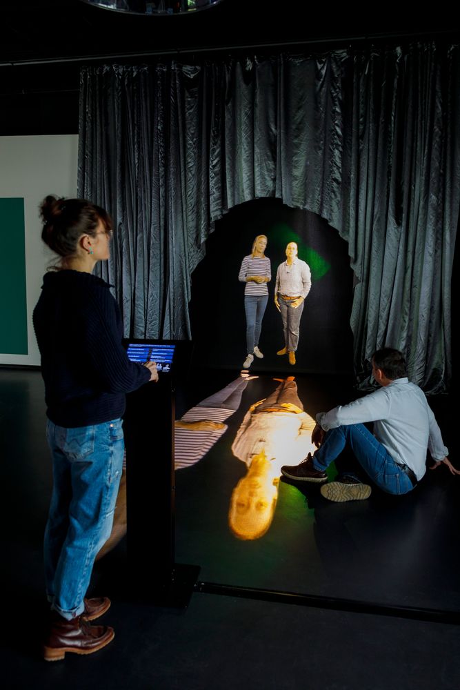 Die Ausstellungsansicht zeigt zwei Personen, die sich ein Hologramm ansehen. Das Hologramm zeigt zwei Frauen, die nebeneinander stehen.
