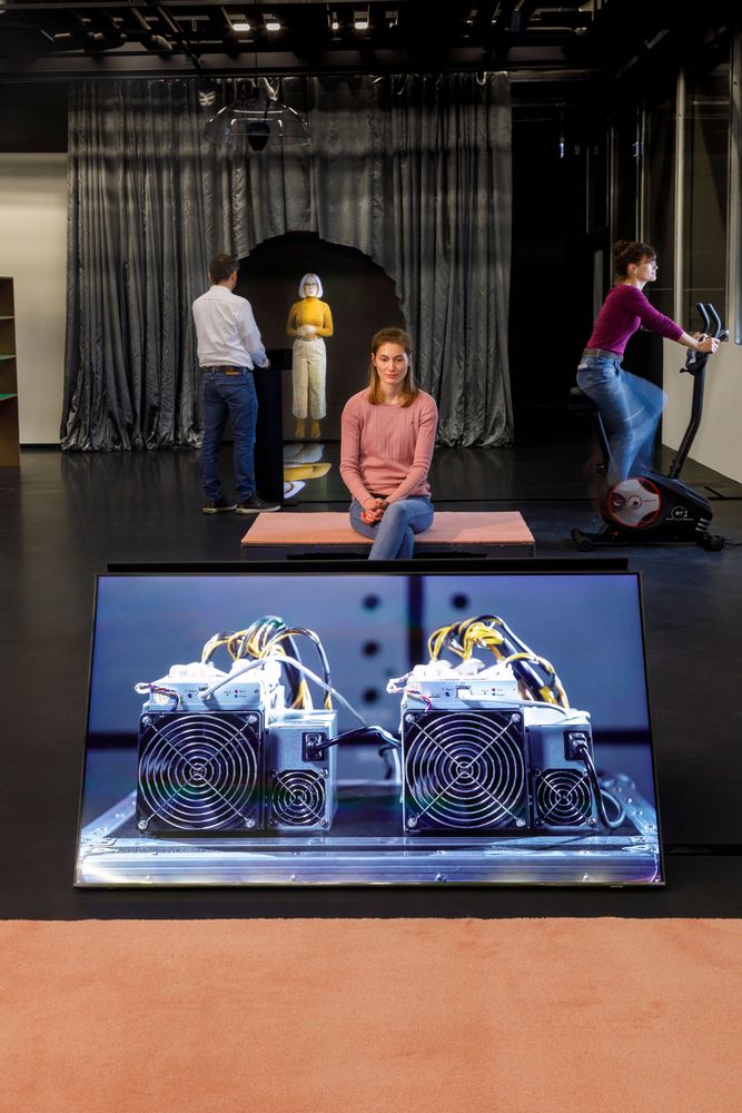 Die Ausstellungsansicht einen Bildschirm, auf dem ein Video abspielt. Dahinter sitzt eine Frau auf und betrachtet den Bildschirm auf der gegenüberliegenden Seite. Rechts sitzt eine Frau auf einem Hometrainer und ein Mann betrachtet ein Hologramm.