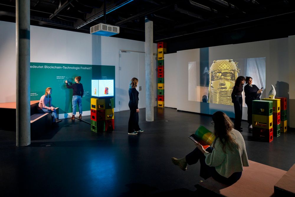 Die Ausstellungsansicht zeigt mehrere Personen die sich in einem Raum befinden. Eine Frau schreibt mit Kreide an eine grüne Wand. Zwei Frauen stehen rechts im Bild vor einem Podest und betrachten eine Statue.