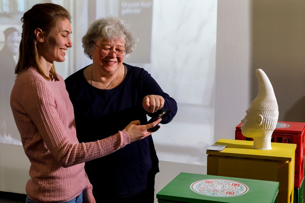 Die Ausstellungsansicht zeigt zwei Frauen, die vor einem Podest mit einer Skulptur stehen und einen QR-Code mit dem Handy einscannen. 