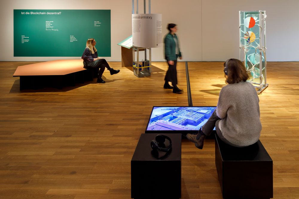 Die Ausstellungsansicht zeigt eine Frau die Kopfhörer trägt und vor einem Bildschirm sitzt. Eine weitere Frau liest einen Ausstellungstext, der an einer Säule befestigt ist. 