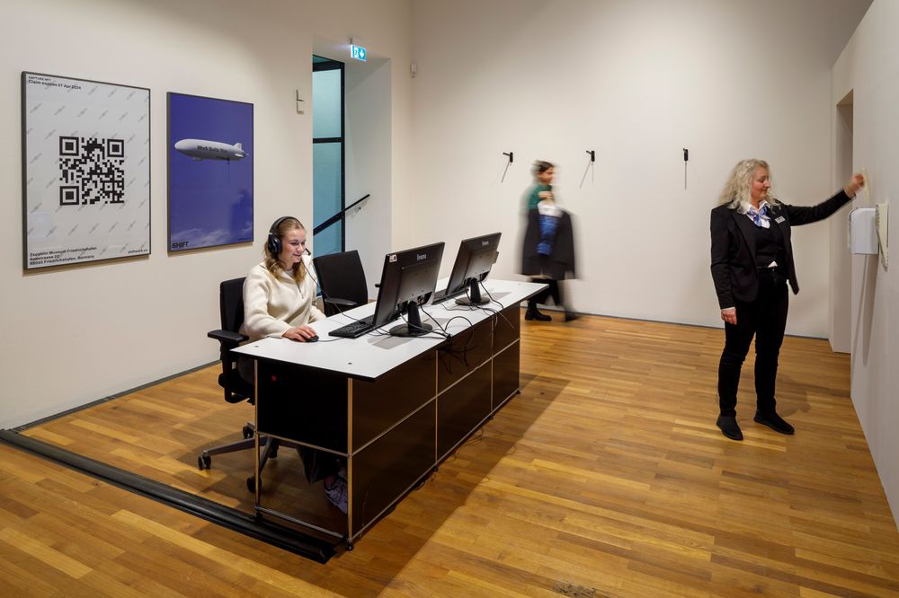 Die Ausstellungsansicht zeigt ein nachgestelltes Büro. An einem Schreibtisch sitzt eine Frau mit Kopfhörern vor einem Computer. Eine Frau aus dem Aufsichtspersonal, die ihre Uniform trägt, stempelt mit einer Karte an einer Stempeluhr ein.
