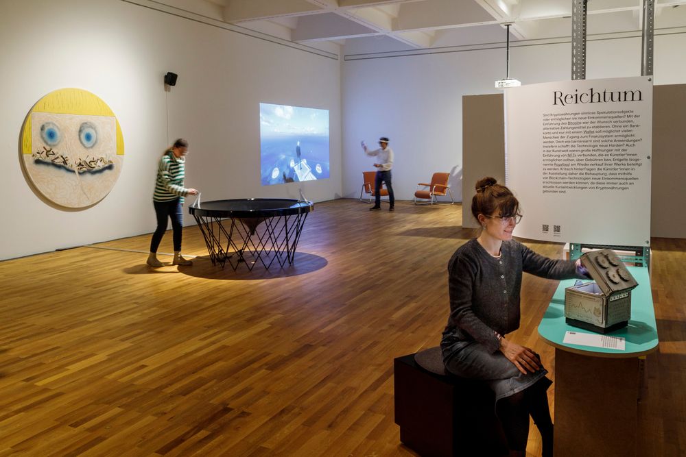 Die Ausstellungsansicht zeigt im Vordergrund eine Frau, die eine Rätselbox öffnet. Im HIntergrund wirft eine Frau eine Münze in eine Installation, die an einen Trichter erinnert. Im Hintergrund steht ein Mann der eine VR-Brille trägt. 