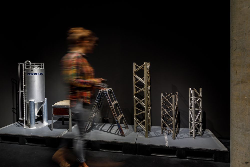 Die Silhouette einer Frau bewegt sich an verschiedenen Ausstellungsobjekten aus Aluminium vorbei. Darunter sind ein Hocker und eine kleine Leiter.
