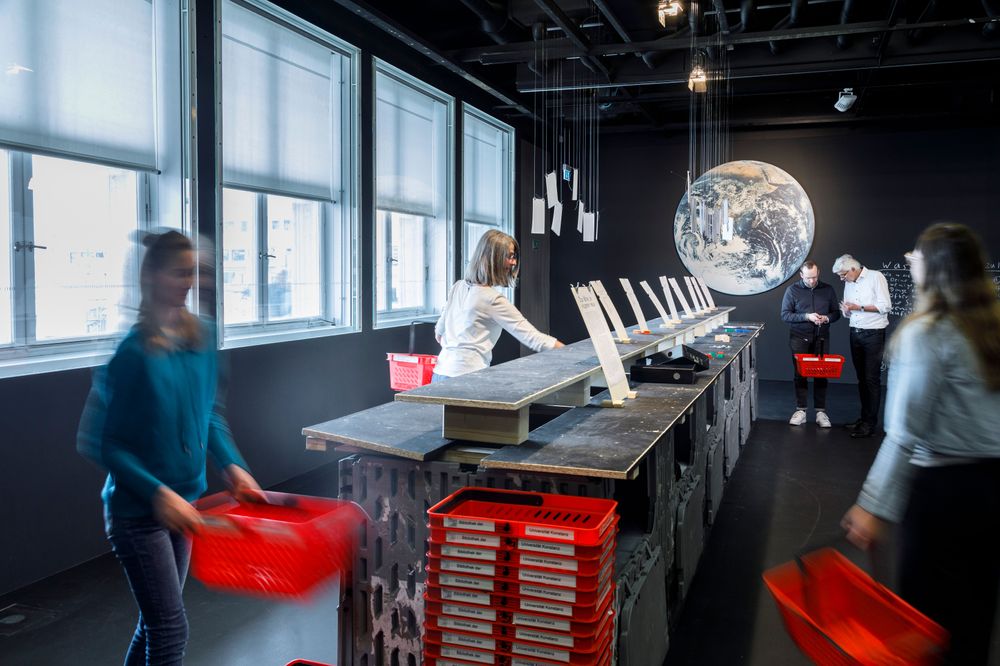 Die Ausstellungsansicht zeigt  mehrere Personen, die sich mit roten Einkaufskörben durch den Raum bewegen. In der Mitte befindet sich eine Theke, aus der man an beiden Seiten verschiedene Objekte greifen kann. 